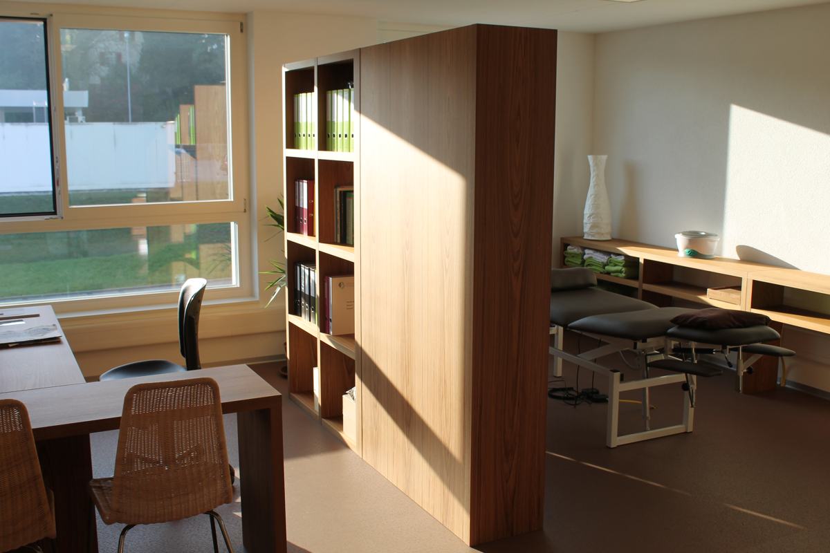 Besprechungstisch und Raumteiler aus Holz, dahinter der Behandlungsraum